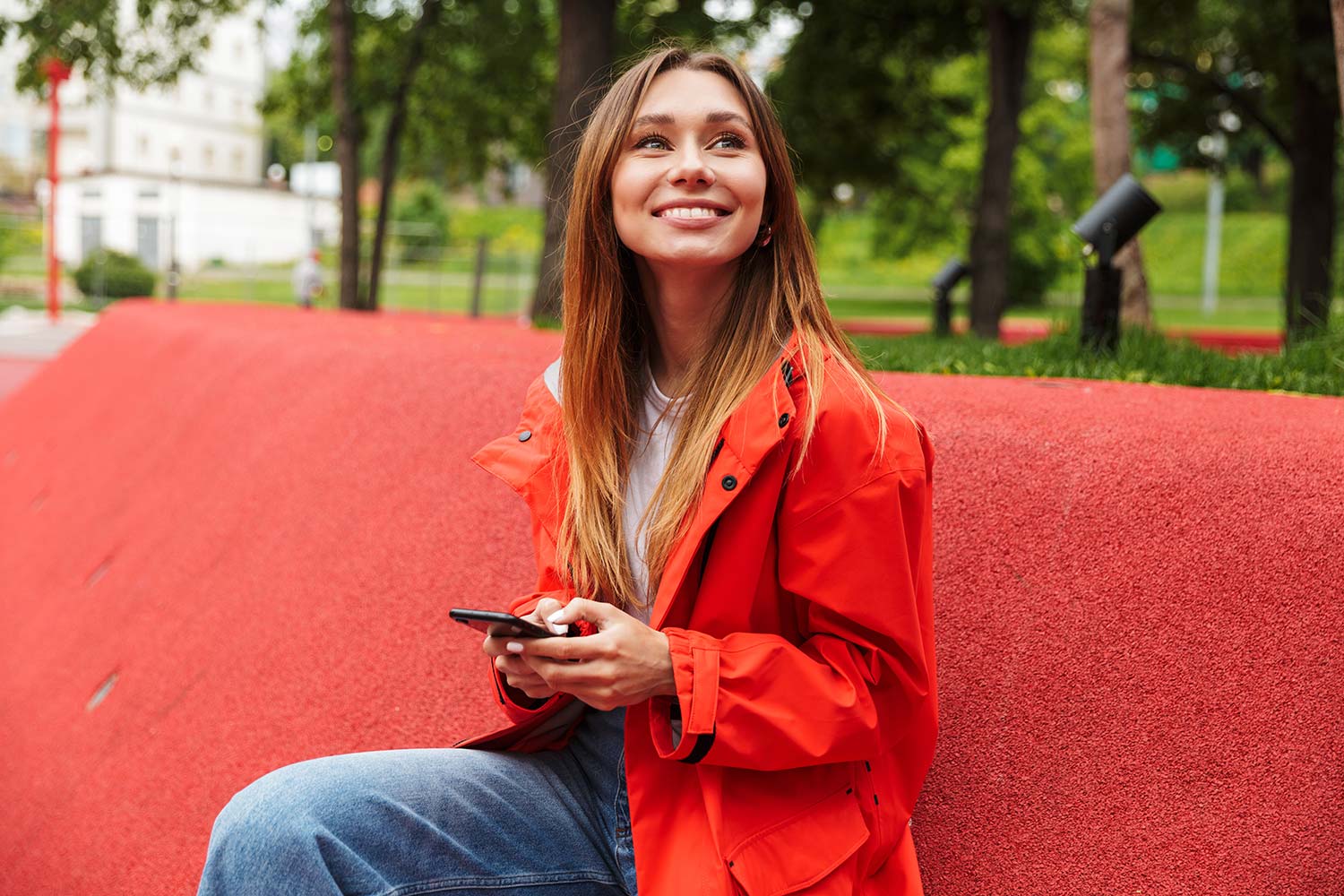 Attraktive lächelnde junge Frau in roter Regenjacke mit Smartphone