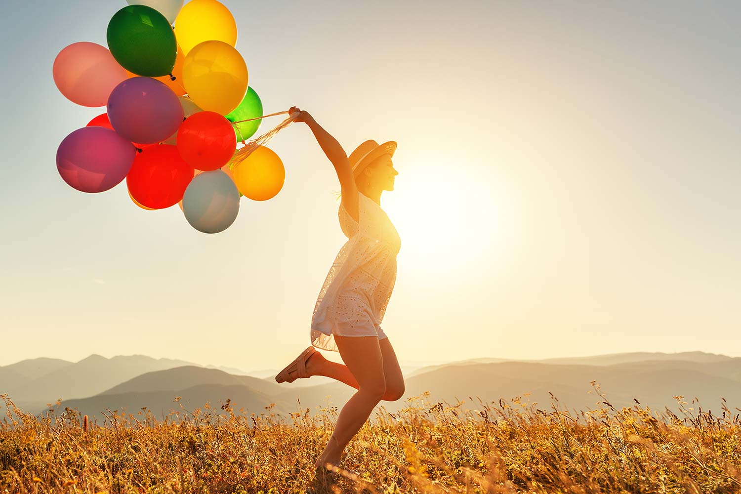 Sommerlich gekleidete Frau mit Luftballons in hügeliger Landschaft bei Sonnenuntergang