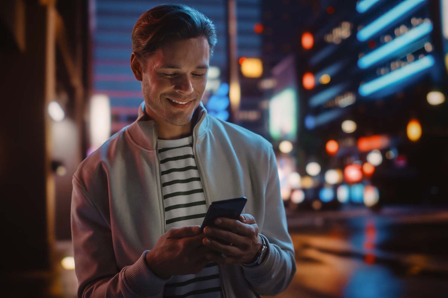 Junger attraktiver Mann mit Smartphone vor nächtlicher, leuchtender Stadtkulisse