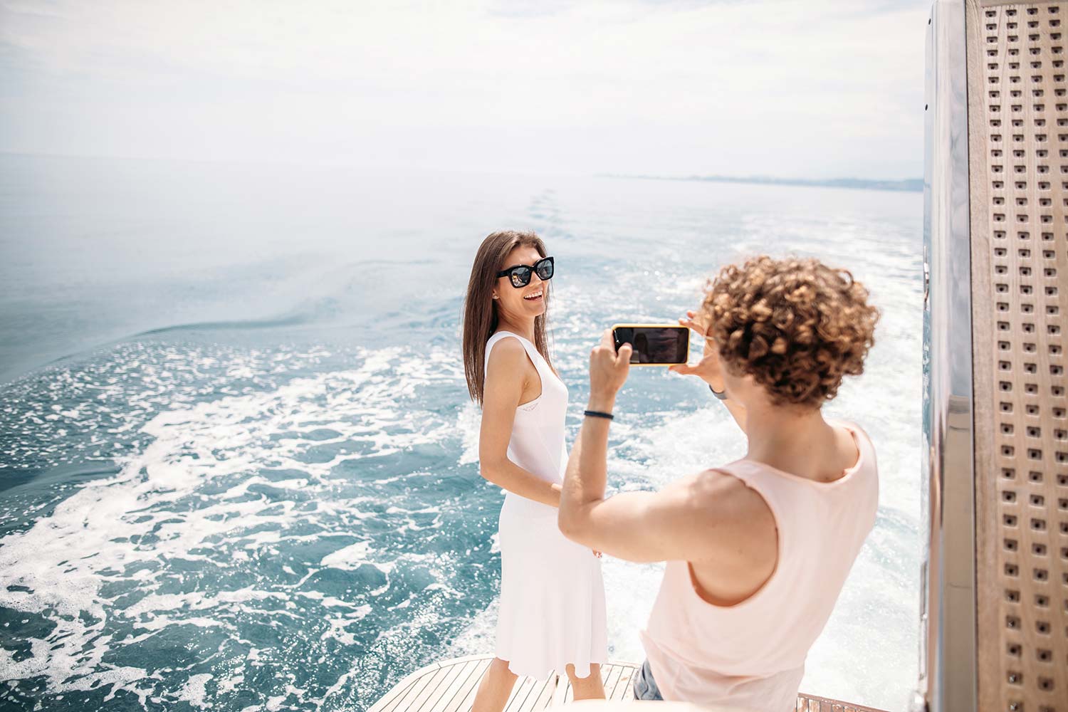 Junger Mann fotografiert Frau in weißem Kleid auf einer Yacht